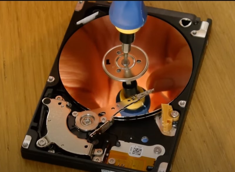 An open hard drive with a blue-handled screwdriver touching its central spindle. The reflective disk inside shows a reddish-orange hue